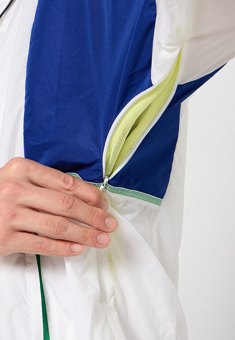 Veste blanche et bleue avec fermeture éclair, dotée d'une doublure en mesh jaune. Main tenant la fermeture éclair, mettant en valeur la texture du tissu de la veste.