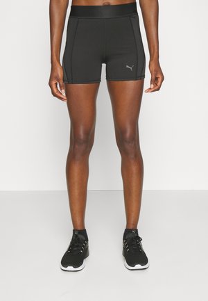 Fit person iført sorte Puma træningsshorts og sorte Puma sneakers stående på et hvidt gulv mod en ensfarvet hvid baggrund.