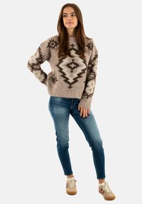 Beige knitted sweater with dark brown geometric patterns, high neckline, ribbed hem. Paired with slim-fit blue jeans and beige sneakers.