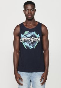 Tumed sinine tank top, millel on graafiline print palmilehtedega ja tekst "Ohara Beach", valmistatud kergest materjalist, kantud heledate teksashortsidega.