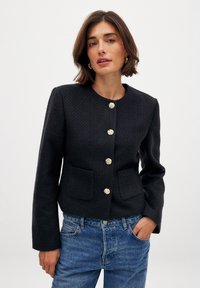 Giacca nera con texture, colletto rotondo, quattro bottoni dorati e due tasche frontali. Indossata con jeans blu.