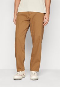 Tan cotton trousers with a relaxed fit, featuring side pockets and a smooth texture, paired with white sneakers.