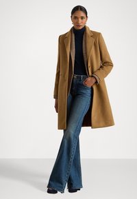 Barna gyapjú külső kabát, benne kivágott gallérral, turtleneck pulóver és kockás zakó fölött viselve, kék, harangszárú farmernadrággal párosítva.