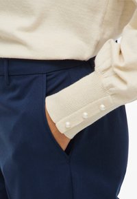 Beežist kootud kampsun koos pärlikujuliste nööpide aktsentidega varrukaserval, mida on kombineeritud tume mereväe toonides õmmeldud pükstega. Sile tekstuur ja kehasse sobiv lõige.