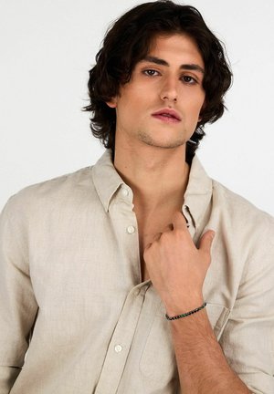 Camicia di lino beige chiaro con colletto abbottonato e una tasca. Un braccialetto di perline con accenti neri e teal adorna il polso.