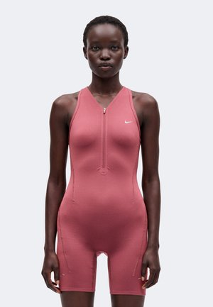Donna in piedi che indossa una tuta atletica rosa Nike senza maniche con zip frontale e pantaloncini su uno sfondo chiaro e uniforme.