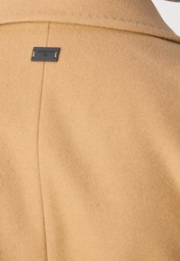 Cappotto color cammello con una texture liscia simile alla lana, caratterizzato da una cucitura posteriore cucita e da una piccola etichetta nera del marchio vicino al colletto.