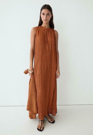 Femme portant une robe maxi plissée sans manches de couleur rouille, tenant un vêtement texturé assorti et portant des sandales noires.