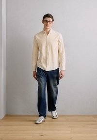 Light beige button-up shirt, short sleeves, denim jeans with a faded effect, and white sneakers with green accents. Model standing against a plain wall.