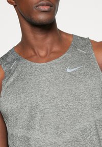 Szary sportowy top bez rękawów wykonany z teksturowanej tkaniny, z okrągłym dekoltem, tonalnym przeszyciem i małym, odblaskowym logo Nike na klatce piersiowej.