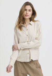 Mujer de cabello largo con una camisa beige abotonada y pantalones marrones claros, de pie frente a un fondo blanco.