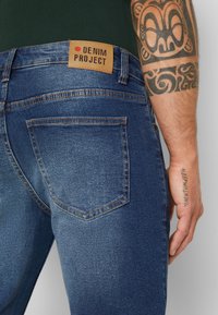 Sötétkék farmernadrág bőr címkével, amelyen az áll "DENIM PROJECT." Hátsó zsebei narancssárga varrással rendelkeznek, és sima tapintásúak.