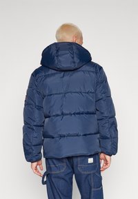 Polstret marineblå hættejakke med vandret syninger, høj krave og elastiske manchetter. Bæres med blå denim bukser med lommer.