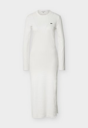 Femme portant une longue robe blanche avec une fente latérale, des chaussettes blanches, des baskets blanches et une casquette noire, debout devant un fond clair uni.
