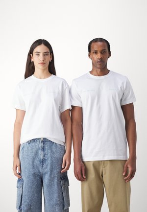 T-shirt in cotone bianco a maniche corte, con un sottile testo stampato in blu nella zona sinistra del petto, abbinato a pantaloni cargo azzurri e pantaloni beige.