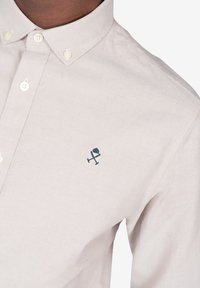 Camisa de botón color beige claro con cuello, que presenta un pequeño logo bordado en azul marino en el pecho y una textura suave de algodón.