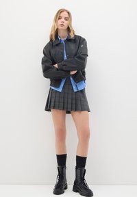 Giovane donna che indossa una giacca bomber nera, una camicia a righe blu, una gonna plissettata grigia, calze nere e stivaletti neri, in piedi contro un muro bianco.