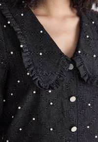 Abito in denim nero con colletto arricciato, decorazioni in perle e un motivo trapuntato. Sono visibili dettagli con bottoni e tessuto testurizzato.