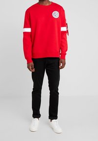 Felpa rossa con collo rotondo, strisce bianche sulle maniche e una toppa con logo. Abbinata a jeans neri e sneakers bianche.