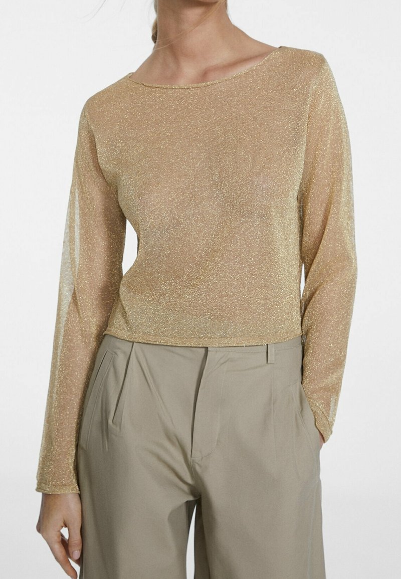 Genomskinlig långärmad topp i metallisk guld med båtformad halsringning, som har en figurnära silhuett och mjuk glittrig textur. Matchas med beige byxor.