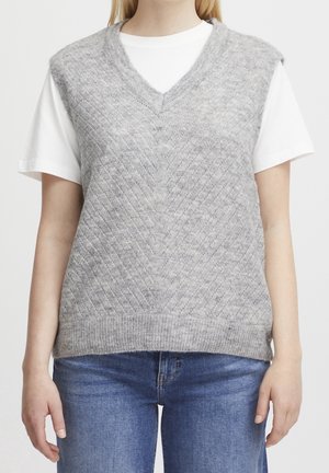 Kobieta nosząca szarą, dzianą kamizelkę bez rękawów z dekoltem w kształcie litery V na białym krótkim rękawem t-shircie i niebieskich dżinsach, widoczna od szyi do bioder.