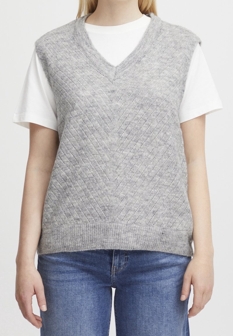 Femme portant un gilet sans manches en laine gris à col en V sur un t-shirt blanc à manches courtes et un jean bleu, visible du cou aux hanches.