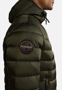 Giacca puffa verde oliva con cappuccio, dotata di sezioni trapuntate, una tasca con zip laterale e una patch circolare del logo sulla spalla.