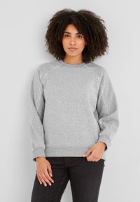 Woman with curly hair wearing a light grey sweatshirt with white stitched seams and black pants, standing against a plain light background.