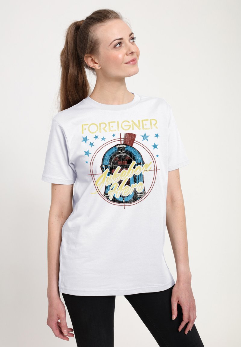 Vit bomull t-shirt med en grafisk design som har blå och gula texter som säger "FOREIGNER Jukebox Hero", omgiven av blå stjärnor.