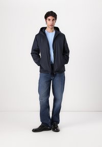 Marineblauwe lichtgewicht hoodie met rits, gecombineerd met een lichtblauw t-shirt, versleten spijkerbroek en zwarte schoenen. Effen witte achtergrond.