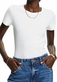 T-shirt bianco a maniche corte, design fitted, morbido misto cotone, indossato con jeans blu, con una collana d'oro visibile.