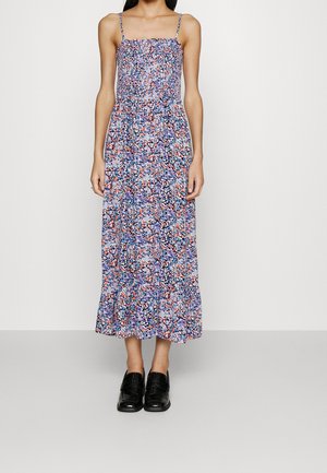 Femme portant une robe fleurie sans manches, longue jusqu'aux chevilles, avec des fines bretelles et des mocassins noirs, debout devant un fond blanc uni.