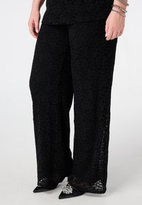 Zwarte wijduitlopende broek van kant met bloemenpatronen, semi-transparante textuur en een comfortabele pasvorm, gecombineerd met zwarte puntige hakken en metallic accenten.