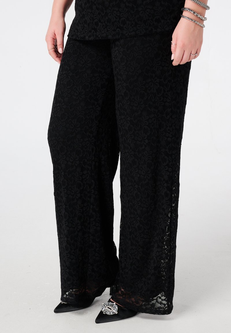 Zwarte wijduitlopende broek van kant met bloemenpatronen, semi-transparante textuur en een comfortabele pasvorm, gecombineerd met zwarte puntige hakken en metallic accenten.