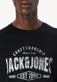 Black cotton T-shirt featuring a large white graphic print with "JACK & JONES" text and vintage-style accents. Crew neck design.