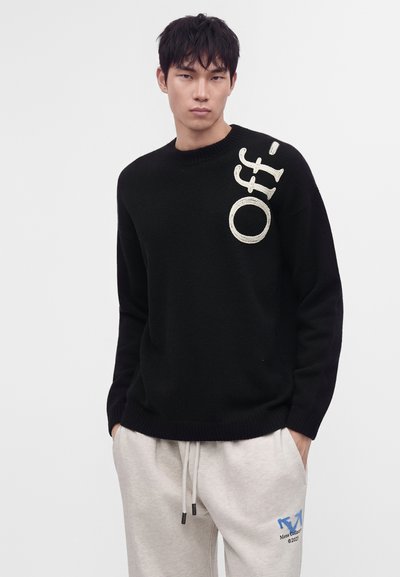 Jeune homme portant un pull noir avec le texte "Off-" sur l'épaule et un pantalon de survêtement gris clair avec impression bleue, debout devant un fond uni.