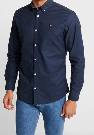 Homme portant une chemise ajustée bleu marine à manches longues et boutons, avec un petit logo rouge et blanc sur la poitrine, associée à un jean bleu clair.