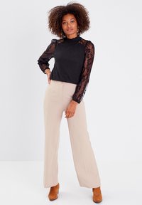Haut noir à manches longues en dentelle, col montant et coupe ajustée. Pantalons évasés beige clair et bottes marron à la cheville complètent l'ensemble.