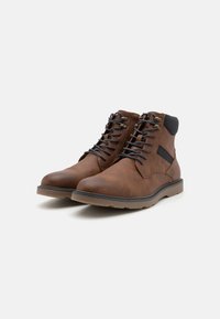 Botas de cuero marrón hasta el tobillo con una textura suave, que presentan un diseño de cordones, ojales de metal y un acento negro contrastante en el lateral.