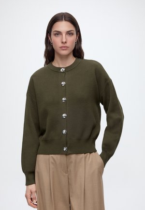 Vrouw met lang bruin haar draagt een olijfgroen gebreid vest met knopen en beige geplooide broek, staand met één hand in de zak tegen een witte achtergrond.