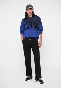 Kék kötött pulóver fekete "VERSACE JEAN COUTURE" felirattal, fekete nadrággal és fekete sportcipővel párosítva, fehér kiegészítőkkel.