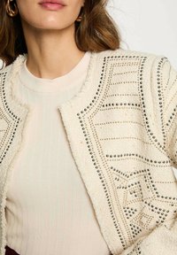 Giacca in tweed color crema con borchie metalliche, linee strutturate e finitura texturizzata. Indossata sopra una camicia a coste leggera.