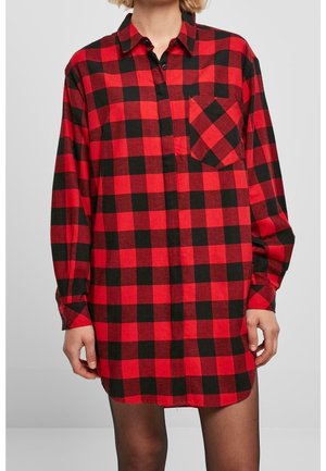 Kobieta w dużej, czerwono-czarnej koszuli w kratę, zapinanej na guziki, z kieszenią, stojąca z rękami swobodnie opuszczonymi wzdłuż ciała.