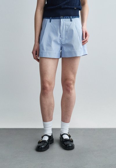 Personne portant un short à rayures verticales bleues et blanches, des chaussettes blanches, des mocassins noirs en cuir, et un haut sans manches marine, se tenant sur un sol gris.