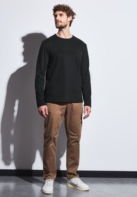 Czarna koszulka z długim rękawem i okrągłym dekoltem, w zestawie z beżowymi chinosami i białymi trampkami, stojąca na tle jednolitej ściany. Widoczny cień.