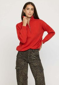 Un pull zippé rouge avec un col montant, associé à un pantalon cargo à imprimé léopard olive, doté de poches latérales et d'un tissu texturé.