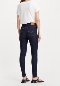 Tmavě modré skinny džíny s vysokým pasem, vyrobené z denimové látky, mají koženou nášivku na zadní straně a kontrastní švy. Jsou kombinovány s bílým tričkem.