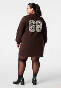 Bruine sweatjurk met witte grafische "68" op de achterkant, lange mouwen, zijsplitten en gecombineerd met zwarte enkellaarzen.