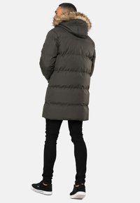 Groene pufferjas met een capuchon en een nepleren rand, verticale gestikte ontwerp en een lengte tot boven de dij. Draagt met zwarte skinny jeans en sneakers.