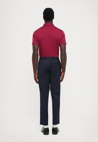 Uomo di spalle che indossa una polo a maniche corte bordeaux, pantaloni a righe blu marino, calzini bianchi e scarpe eleganti nere su uno sfondo semplice.